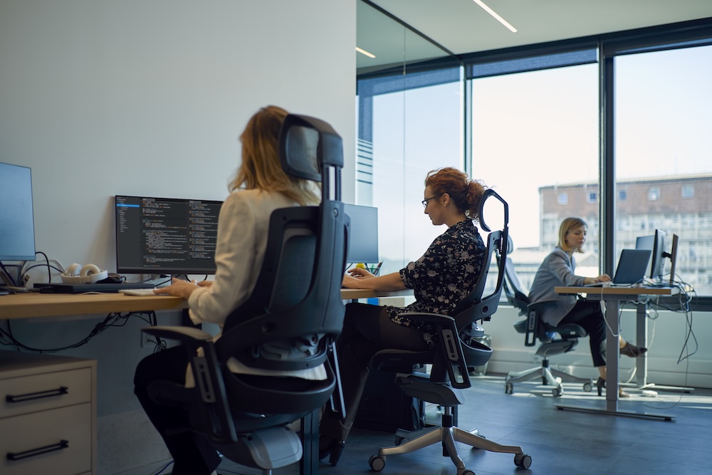 Prevenirea afecțiunilor musculoscheletice într-un mediu ergonomic Angajate lucrând la birouri ergonomice într-un mediu modern, ilustrând prevenirea riscurilor musculoscheletice la locul de muncă.